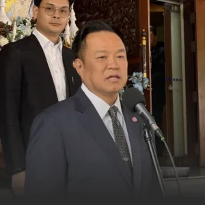 A man in a dark suit and tie speaks into a microphone at a press briefing, with another suited man standing behind him near a decorative doorway.