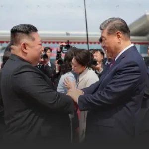 Two men in suits shake hands at a public event, with photographers and onlookers in the background.