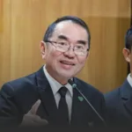 Smiling man in a dark suit and tie speaks into a microphone at a formal event.