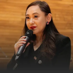 Woman in a black blazer speaks into a handheld microphone against a wooden backdrop.