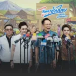 Thai officials at a press conference with many colorful microphones in front of a market-themed backdrop.