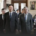 Two suited men walking side by side in a formal hallway, with others in suits following behind in a professional setting