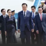 Group of formally dressed officials walking off an airplane, led by a man in a navy suit and striped tie, with a woman in a dark polka-dot dress beside him, others in suits follow.