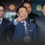 Asian man in a navy suit points toward the audience while speaking at a podium with a seal, flanked by aides in formal attire.