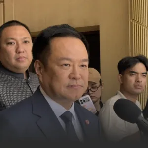 อนุทินย้ำปม นพสรณ พ้นประธาน กสทช ต้องดำเนินการตามกฎหมาย » The Mainstream Man in a dark suit and tie speaks to reporters outside a building, with microphones in front and photographers in the background.