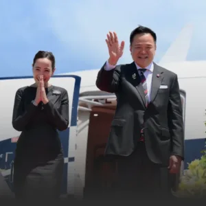 Man in a dark suit waves at the camera beside an airplane as a woman in a black dress stands with hands pressed together in greeting.