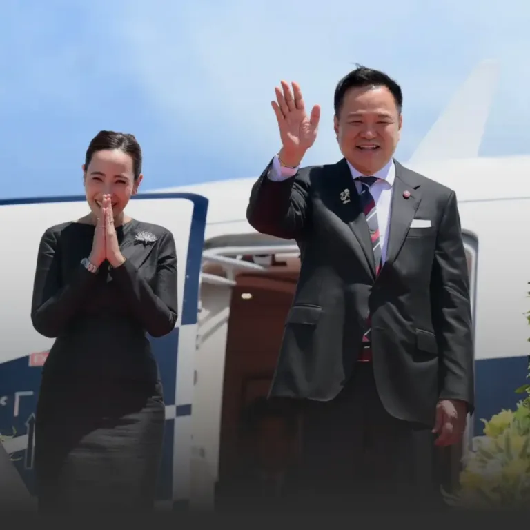 Man in a dark suit waves at the camera beside an airplane as a woman in a black dress stands with hands pressed together in greeting.