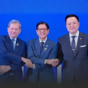 Three men in dark suits shake hands in a row against a blue backdrop, smiling and wearing lapel pins.