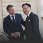 Two men in dark suits shake hands and smile outdoors in front of a stone building, signaling a formal meeting or greeting.