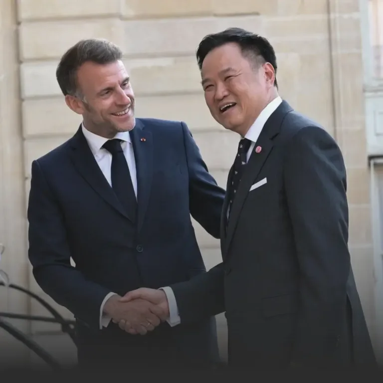 Two men in dark suits shake hands and smile outdoors in front of a stone building, signaling a formal meeting or greeting.