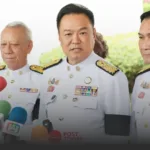 Senior official in a white ceremonial uniform with medals, speaking to reporters at a press conference with microphones in view.