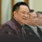 Smiling man in a maroon patterned shirt seated at a conference table, with colleagues in the background.