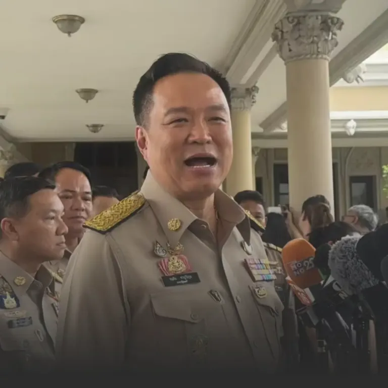 Government official in a beige uniform speaks to reporters, microphones in front, columned building in the background.