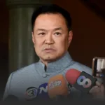 Man in a light blue mandarin-collar shirt speaks to reporters at a press conference, with microphones in front of him.