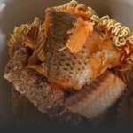 Fried fish fillet with crispy skin resting atop a breaded crust and curly ramen noodles in a reddish sauce on a plate.
