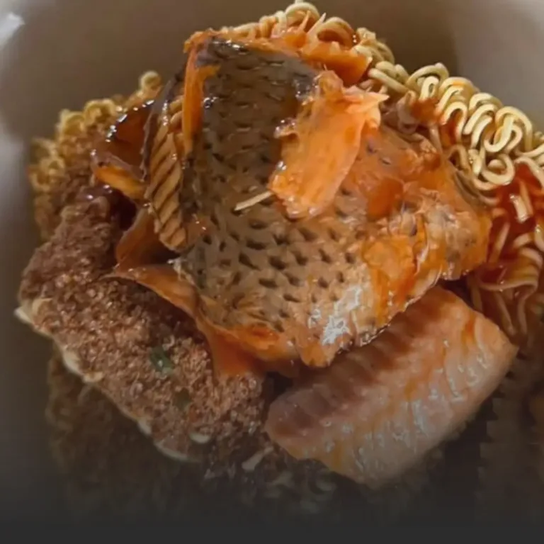 Fried fish fillet with crispy skin resting atop a breaded crust and curly ramen noodles in a reddish sauce on a plate.