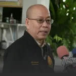 Man in glasses being interviewed, speaking to reporters with colorful microphones in a lobby or indoor garden setting.