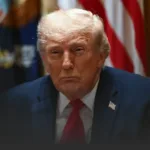 Close-up of a man with light hair in a navy suit and red tie, with a U.S. flag in the background.