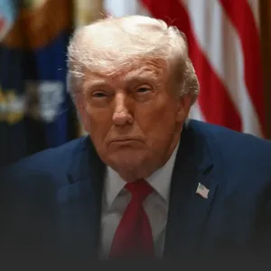 Close-up of a man with light hair in a navy suit and red tie, with a U.S. flag in the background.