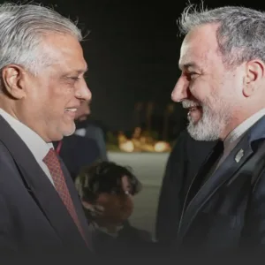 Two older men in dark suits share a smile, facing each other during a nighttime event.