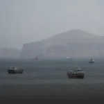 Several large ships floating on a calm sea with a hazy rocky coastline in the background.