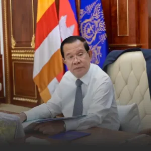 Man in white shirt and tie sits at a desk in a formal office, with flags in the background behind him.