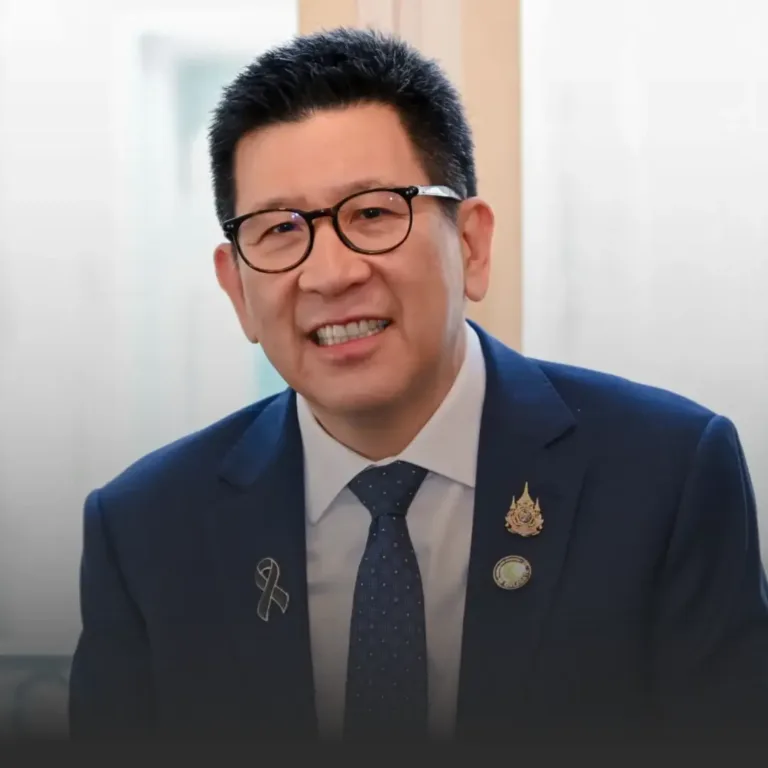Smiling man in a navy suit and glasses, with lapel pins, in a professional indoor setting.