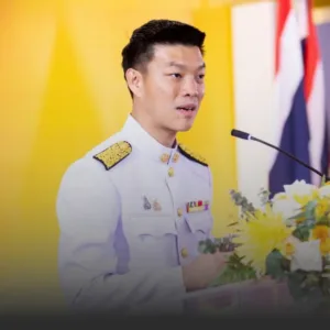 Asian man in a white ceremonial uniform with gold epaulettes speaking at a podium with a microphone and yellow backdrop, floral arrangement nearby.