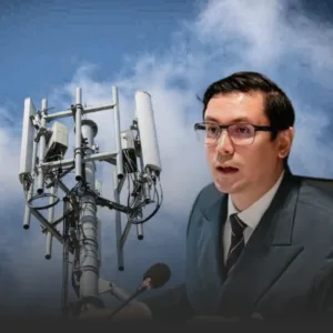 Man in a suit speaks into a microphone beside a large cell tower against a cloudy sky.