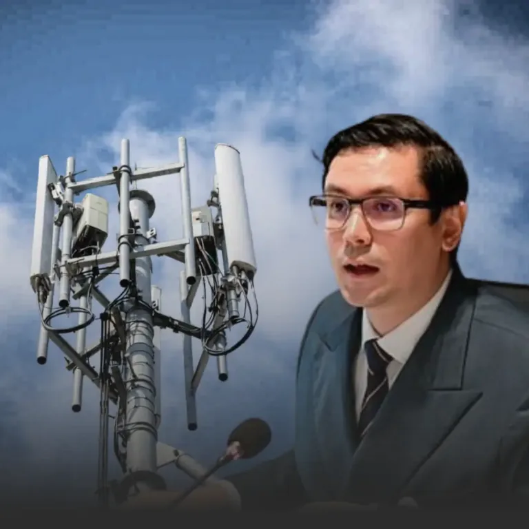 Man in a suit speaks into a microphone beside a large cell tower against a cloudy sky.