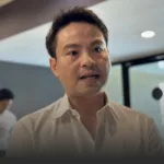 Close-up of a man in a white shirt speaking, indoors in a modern office setting with glass walls in the background.