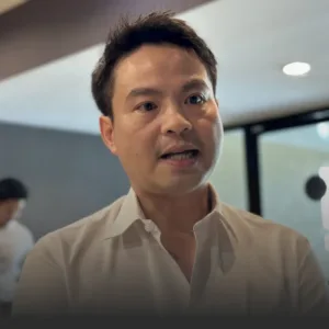 Close-up of a man in a white shirt speaking, indoors in a modern office setting with glass walls in the background.